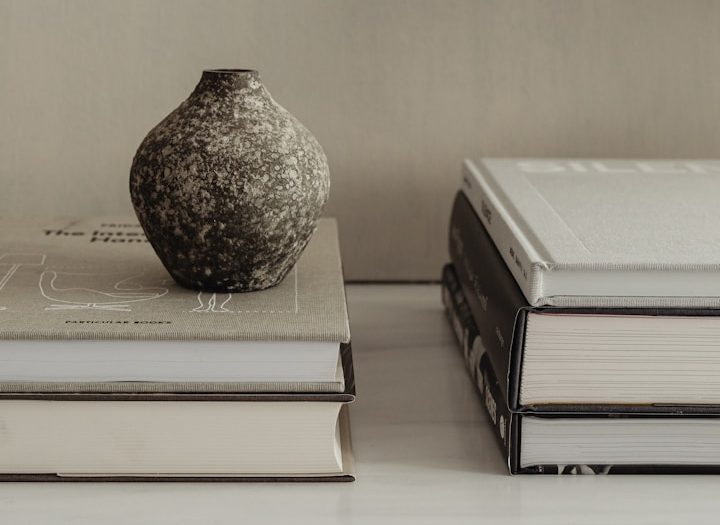 Photo of two stacks of large hard bound books, with a small round black and grey clay vase on top of one stack.